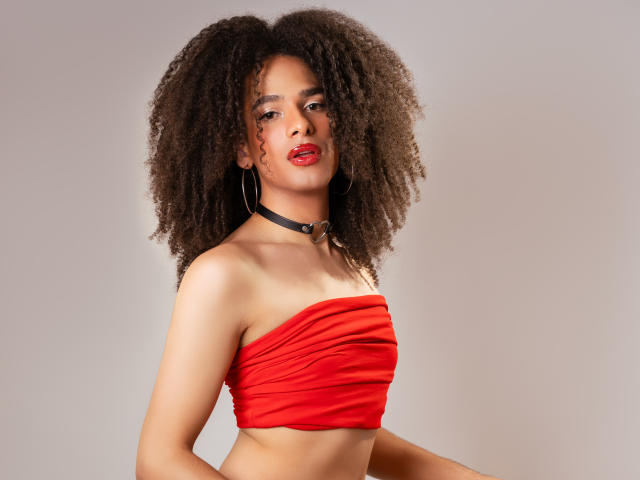KataleyaFowler with big curly brown hair, red strapless top, black choker, red lipstick, looking at camera, showing her tits, hoop earrings, medium skin, confident, sexy, studio shot.