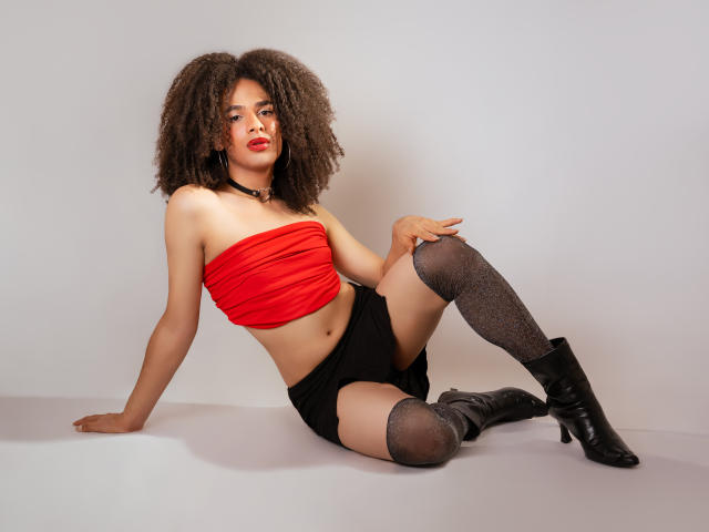 KataleyaFowler with curly brown hair, red tube top, black shorts, gray thigh-high socks, and black boots, sitting with one leg up, showing her ass and a confident look.