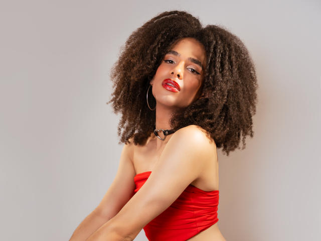 KataleyaFowler with big, fluffy brown afro, wearing a red strapless top, black choker, and red lipstick, looking slightly to the side with a confident, sexy expression.