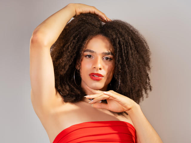 KataleyaFowler with big, curly black hair, red strapless dress, and red lipstick, raising her right arm, showing her armpit, with her left hand near her chin.