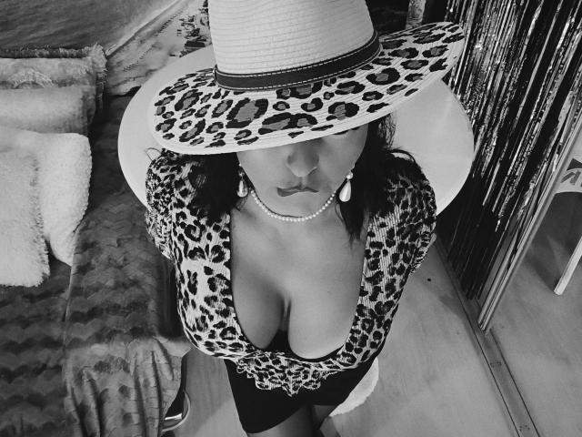 AmberSunshine con una maglietta a stampa leopardo, scollatura profonda, seno prosperoso, indossa un cappello a leopard, una collana di perle e orecchini, guarda verso l'alto, il cappello ombreggia il suo viso, foto in bianco e nero..