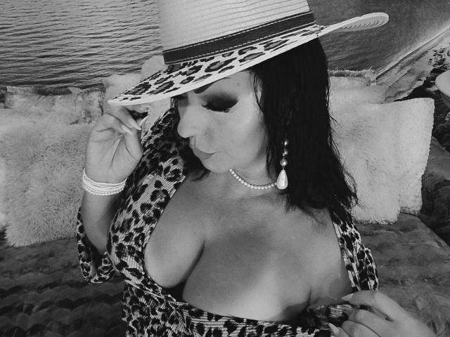 AmberSunshine in een jurk met luipaardprint, die een hoed vasthoudt, haar grote borsten laat zien, een parelketting draagt en oorbellen, met een schattende glimlach en gesloten ogen. Zwart-wit foto..