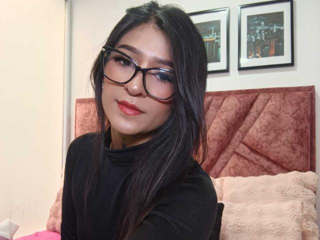 LuciadelaVega has black glasses, straight long hair, red lipstick, and a nose ring, wearing a black turtleneck, sitting on a pink velvet bed, smiling at the camera.