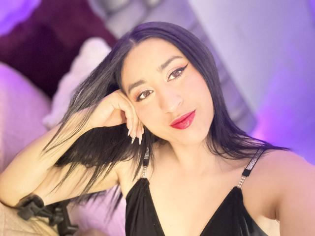 SarahBlakee, met lang zwart haar, rode lippenstift en een zwarte jurk, leunt haar hoofd op haar hand, glimlacht naar de camera en laat haar borsten en schattige gezicht zien..