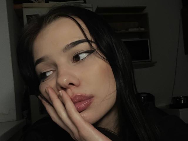 ElvaPaup, avec ses cheveux noirs, sa peau pâle et ses lèvres pulpeuses, une main sur son visage, regardant sur le côté, des yeux sombres, des sourcils épais, des lèvres légèrement entrouvertes de couleur rose, dans une pièce faiblement éclairée..