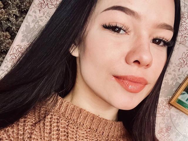 ElvaPaup mit langem, schwarzem Haar, braunen Augen und dicken Wimpern, trägt einen braunen Strickpullover, hat leicht geschürzte Lippen in leuchtendem Orange und ein kaum merkliches Lächeln..