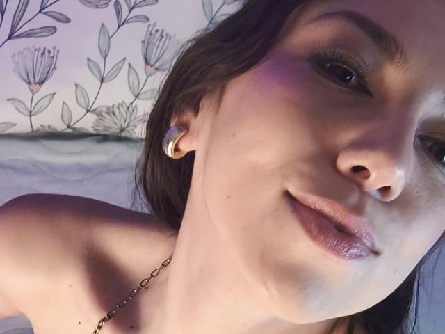 MiaReed's face, showing a slight smile with glossy lips, wearing a gold hoop earring, and a delicate chain necklace, with soft purple light on her right cheek.