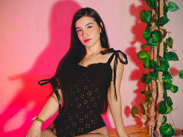Jade Harpper, con lunghi capelli neri, indossa una maglia nera in pizzo con spalline a nastro, pantaloni a rete e un braccialetto dorato, seduta contro un muro rosa con una pianta..