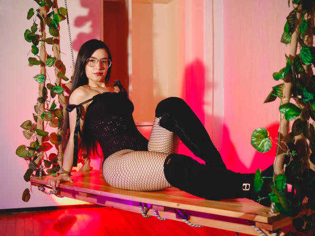 Jade Harpper, con lunghi capelli scuri, occhiali, una maglia nera a spalle scoperte, collant a rete e stivali neri alti fino alla coscia, seduta su un'altalena di legno con rampicanti di foglie, illuminazione rossa..