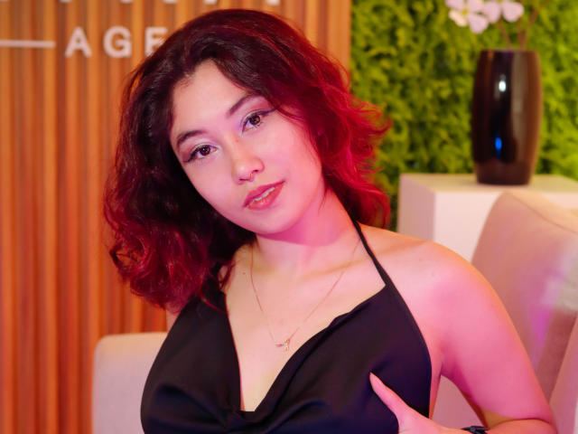 AliCipher, con i capelli rossi ricci, un vestito nero a fascia, seno di media grandezza, pelle chiara e rossetto rosa, indossa una collana d'oro, guarda la fotocamera con un leggero sorriso..