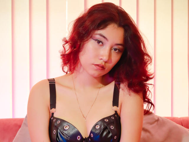 AliCipher, con i capelli rossi ricci, indossa un reggiseno di pelle nera con borchie argentate, guarda in basso con un leggero broncio, ha una collana, la pelle chiara, l'illuminazione è rosa e c'è un divano rosa..
