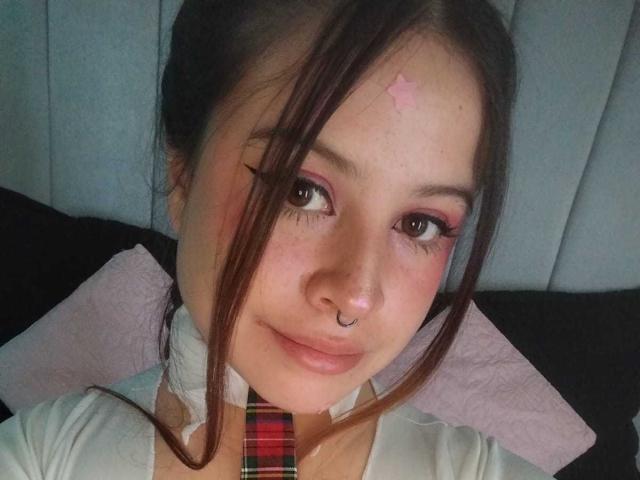 Melani Becker ha un trucco rosa per gli occhi, un anello al naso e una stella rosa sulla fronte. Indossa una cravatta a quadri con una camicia bianca, guardando verso la telecamera..