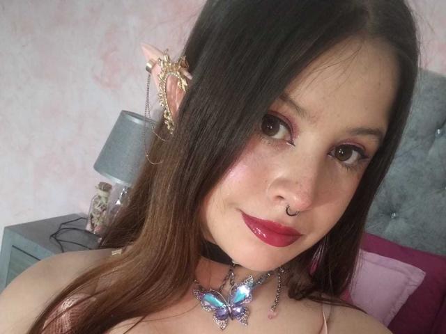 Melani Becker, con i capelli castani, rossetto rosso, piercing al naso e una collana con un ciondolo a forma di farfalla, indossa un collare nero con un fermaglio orecchino decorato, e guarda la fotocamera con un leggero sorriso..