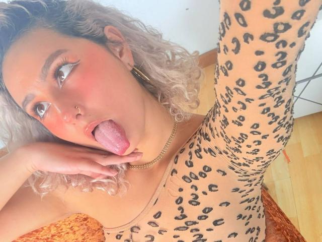 ElenaSaenz with curly blonde hair, leopard print dress, gold hoop earrings, and nose ring, sticking out her tongue with pink tongue and blushed cheeks, wearing a gold chain.