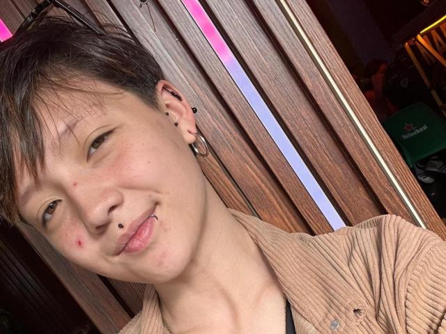 LissyLuxe has short brown hair, a nose piercing, and a lip piercing. She's wearing a beige ribbed shirt, with small zits on her cheek and a slight smirk.