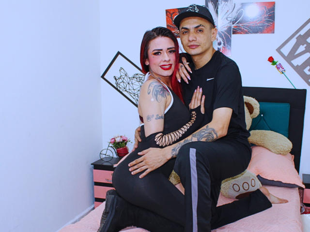 VogueAndPitter with red hair, black sleeveless top, and tattoos, sitting on a bed, ass up, being hugged by VogueAndPitter in black clothes and cap.