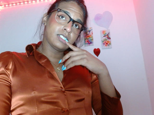 IdiiaLatina with dark skin and glasses, wearing an orange satin shirt, biting her blue-painted fingernail with a playful expression. Her hair is in a loose ponytail.