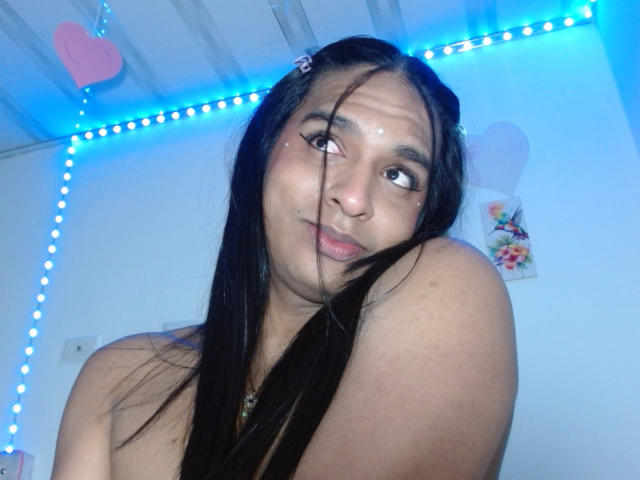 IdiiaLatina with long black hair, looking up with a playful smile, has small tits, a cute face, and a shiny spot on her cheek, in a room with blue LED lights.