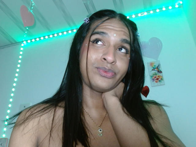IdiiaLatina with long black hair, pouty pink lips, and a gold necklace, holding her chin, looking up, neon blue lights in the background, and a small flower clip.