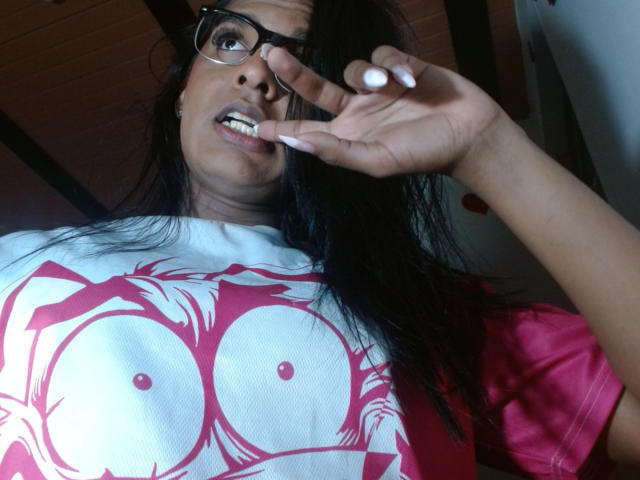 IdiiaLatina with dark hair, glasses, and white-pink shirt showing huge tits, biting nail, looking up, in a low-light indoor setting.