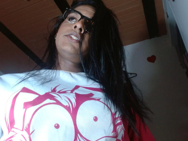 IdiiaLatina with long black hair, wearing glasses, white shirt with red graphic, and looking down at camera, showing her tits. Red heart sticker in background.