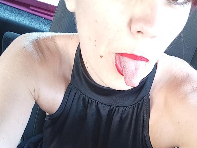 OrianaLaFrancaise, la langue tirée, rouge à lèvres, haut de maillot noir à enfiler, aisselle visible, peau claire et petites taches de rousseur sur la joue, dans une voiture..