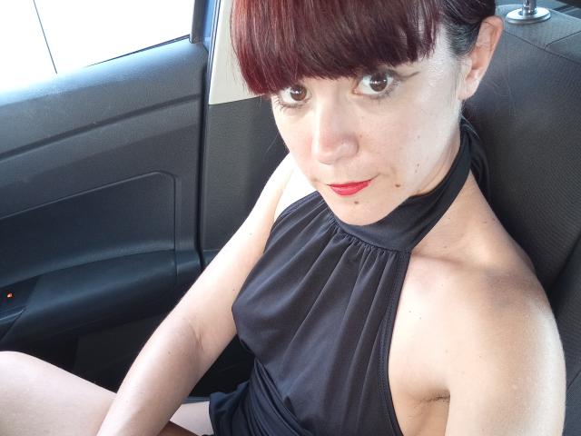 OrianaLaFrancaise has red hair, pale skin, and wears a black halter dress, sitting in a car with a small red light, red lipstick, and a slight smile.