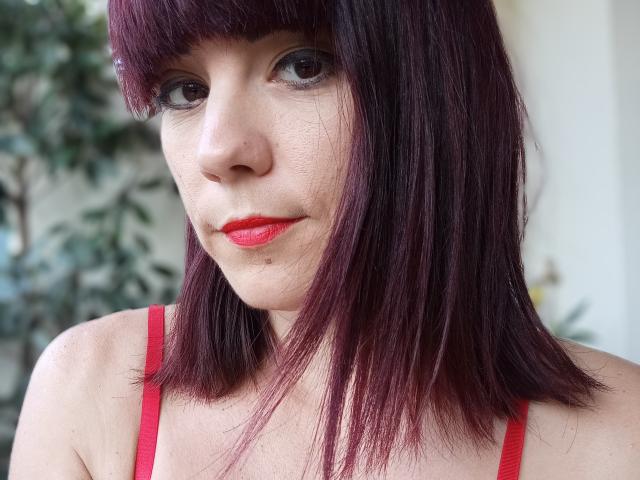 OrianaLaFrancaise, avec une coupe de cheveux courte et droite de couleur violet foncé, du rouge à lèvres rouge et un soutien-gorge rouge, montrant son visage et sa partie supérieure de la poitrine, regardant l'appareil photo avec un léger sourire..