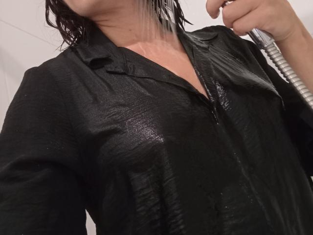 La maglietta nera bagnata di OrianaLaFrancaise aderisce al suo seno, l'acqua scorre sul suo petto, tiene un soffione nella mano destra, capelli scuri e bagnati, sfondo con una parete rivestita di piastrelle bianche..