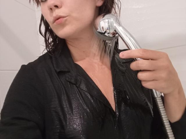 OrianaLaFrancaise avec une chemise noire mouillée, tenant une pomme de douche contre sa poitrine, de l'eau jaillissant, lèvres boudeuses, peau claire, cheveux bruns, seins légèrement visibles, arrière-plan de douche avec des carreaux blancs..