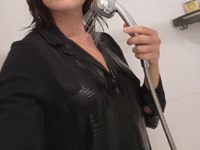 OrianaLaFrancaise con una camicia nera bagnata, che tiene un soffione doccia contro il collo, con l'acqua che schizza, clavicola visibile, capelli scuri corti, mano destra che tiene il soffione, sfondo di parete con piastrelle bianche..