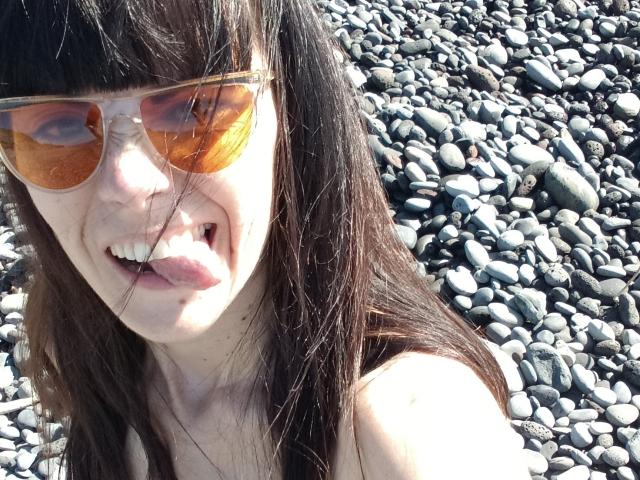 OrianaLaFrancaise, avec de longs cheveux bruns, la langue tirée, portant des lunettes de soleil aviateur orange, les épaules dénudées, debout sur une plage rocheuse..