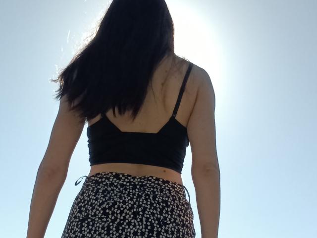 OrianaLaFrancaise ha la schiena rivolta verso la telecamera, mostrando il suo reggiseno sportivo nero e la gonna nera con fiori. La luce del sole crea un effetto alone dietro di lei. Ha i capelli lunghi e scuri..