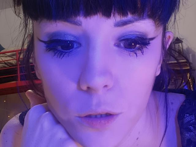 OrianaLaFrancaise, avec un maquillage aux reflets bleus, un eye-liner noir et de longs cils, regardant vers le bas avec une légère moue, la main près de son menton, cheveux noirs avec une frange, teint de peau violet-bleu..