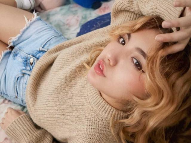 CarmesiLm, con cabello rubio ondulado, vestida con un suéter beige de punto y pantalones cortos vaqueros deshilachados, acostada en una cama, con una mano en el cabello, labios fruncidos, ojos marrones, mirando a la cámara..