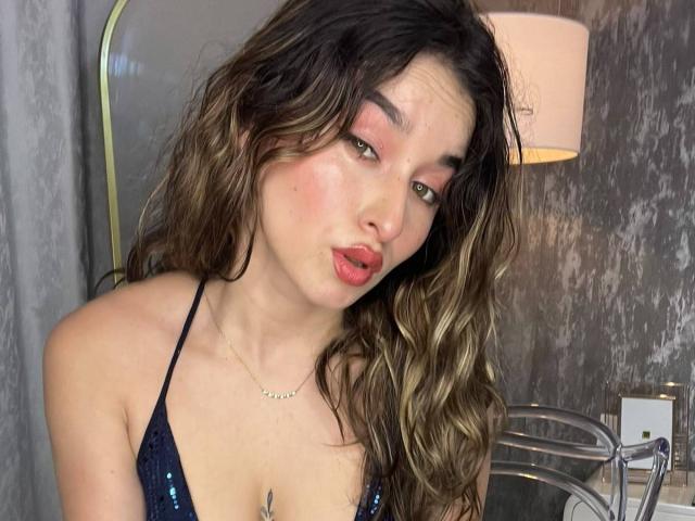 MilaGonz mit welligem, braunem Haar, rosa Lidschatten und rotem Lippenstift, trägt einen glitzernden, schwarzen Bikini-Oberteil und zieht leicht die Lippen zusammen. Sie hat ein süßes Gesicht und eine kleine Brust..