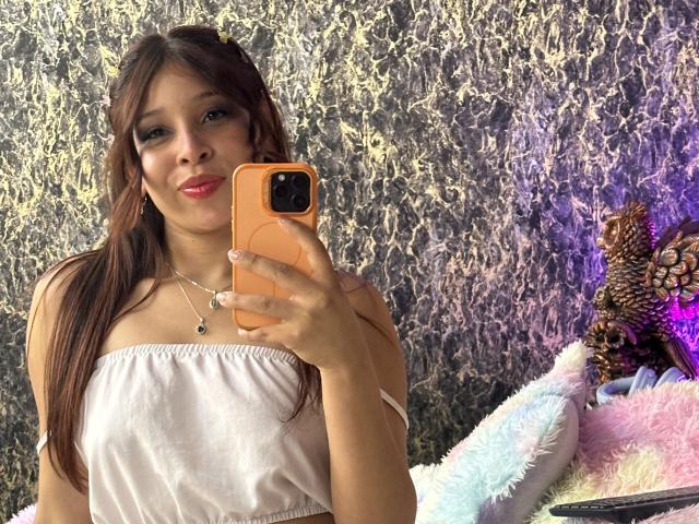 JaneKetty with long brown hair, wearing a pink strapless top, holding an orange phone, smiling, pink lipstick, necklace, pink and white fluffy pillow, pine cone decoration, textured wall.