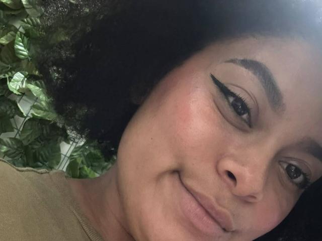 ElizabethBloom's face, dark curly hair, brown skin, black eyeliner wings, subtle smile, pinkish cheeks, and small nose. Her ass isn't visible, just her face and upper chest.