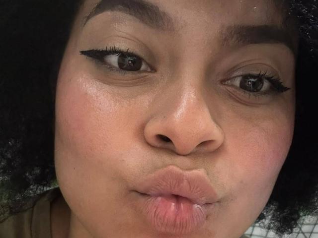 ElizabethBloom's face with dark curly hair, brown eyes, black eyeliner, and pouty pink lips, showing a slight kiss. Her skin is smooth with a light pink blush.