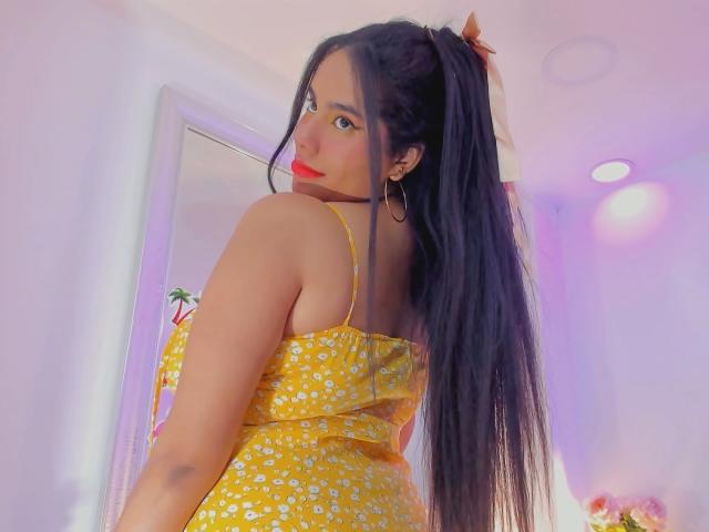 Sienna Taylor, con lunghi capelli neri raccolti in una coda di cavallo, indossa un abito giallo con fiori, guarda indietro con il rossetto rosso, mostrando la schiena e piccoli orecchini a cerchio. Il suo sedere è visibile..