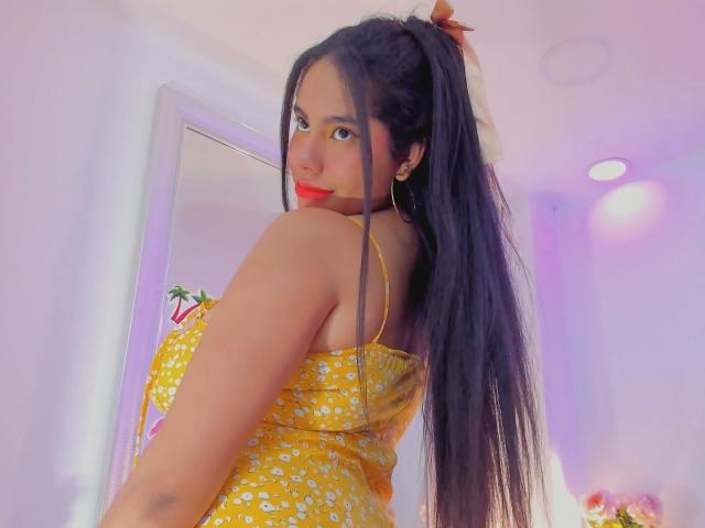 Sienna Taylor mit langem, schwarzem Haar im Pferdeschwanz, gelbem Blumenkleid, rotem Lippenstift, die über die Schulter schaut und dabei ihren Rücken und ihre kleinen Brüste zeigt, in einem hell erleuchteten Raum..