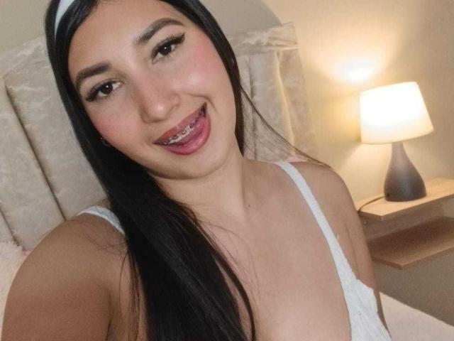 LizzyDilucas with long black hair, wearing a white tank top, sticking out her tongue with metal braces, smiling at the camera, showing her small tits and cute face.