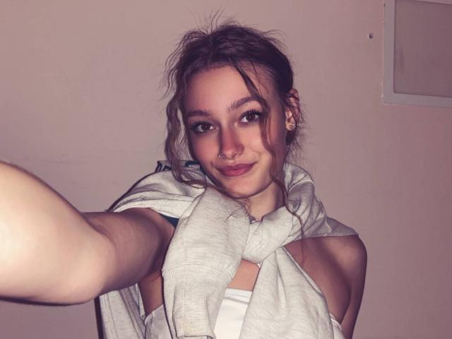 LeilaBie hat zerzaustes braunes Haar, einen Nasenring und ein verschmitztes Lächeln. Sie trägt einen grauen Schal, und ihr weißes Oberteil lässt ihre Brustansätze erkennen. Sie macht ein Selfie..