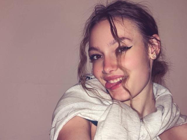 LeilaBie mit zerzaustem, braunem Haar, die ein weißes Hemd trägt, lächelt mit dunklem Eyeliner, hat einen Nasenring und kleine Ohrringe, und hält ein graues Tuch über ihrer Schulter..