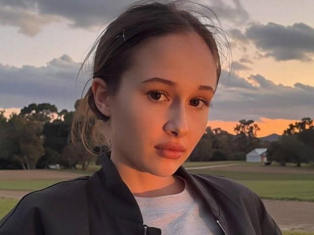 MonaCloudI with pouty lips, brown eyes, and hair in a small clip, wearing a dark jacket over a grey shirt, standing against a sunset sky.