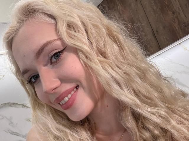 SheraSwayzer's blonde, wavy hair and pale skin, smiling with pink lips and subtle makeup, showing her cute, small nose and blue eyes. Her tits are not visible.