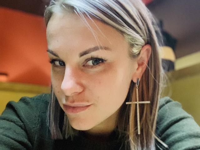 BelleLisaG, con la pelle chiara, gli occhi azzurri e i capelli biondi lisci, indossa una maglietta verde e un orecchino a forma di croce, guarda leggermente di lato con un sorriso appena accennato..