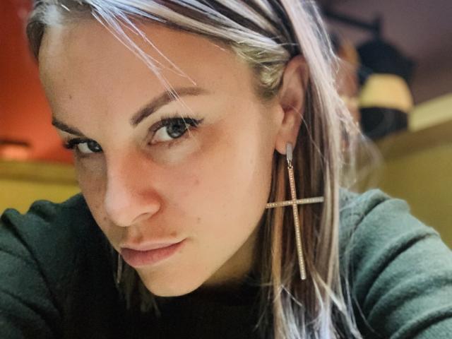 BelleLisaG has blonde hair with white highlights, green eyes, and a serious look. She's wearing a cross earring and a green top, with slight redness on her face.