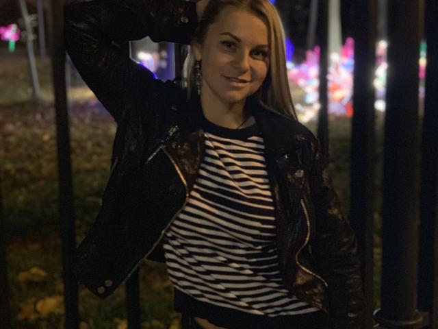 BelleLisaG with blonde hair, wearing a black leather jacket and striped shirt, smiling, hand in hair, dark background with colorful lights, night setting, looking cute and confident.
