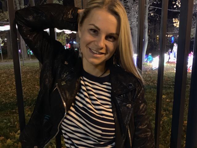 BelleLisaG con i capelli biondi, sorridente, che indossa una giacca di pelle nera e una camicia a righe, con una mano tra i capelli, in piedi vicino a una recinzione metallica di notte, con delle luci sullo sfondo..